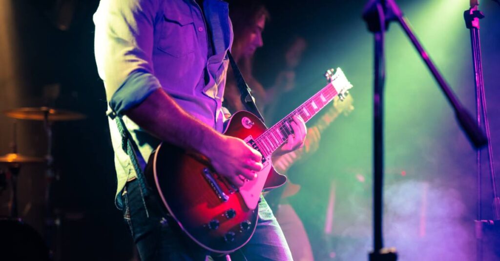 A man stands on a stage, playing a guitar. The stage is darkly lit with fog rising up. A second guitarist is in the background.