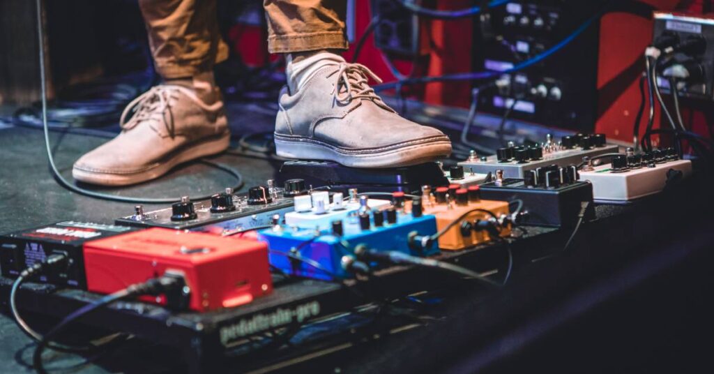 A person is standing in front of a pedalboard with various footswitches attached to it and wires coming off of it.