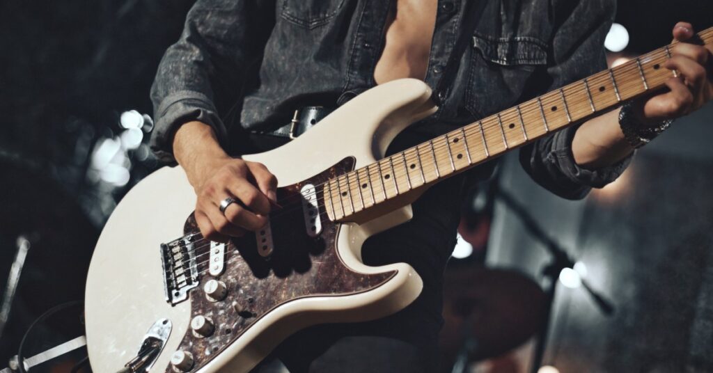 A musician standing on a stage playing a white electric guitar while wearing a jean button-up shirt.