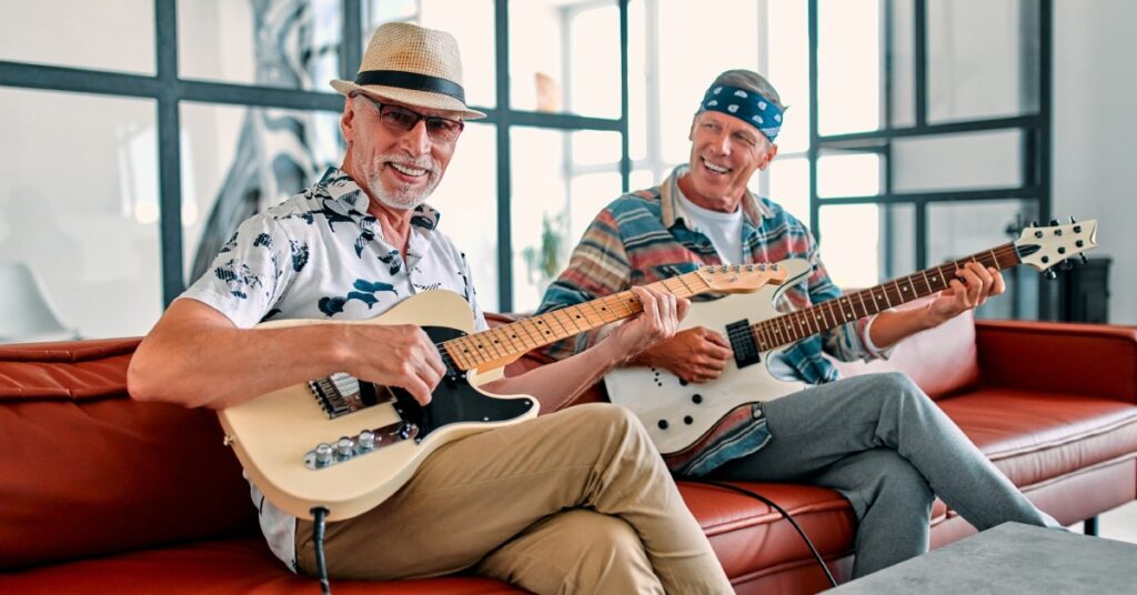 Two men sitting on a red couch smiling and laughing with guitars in their hands in front of a coffee table.